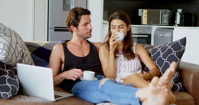 Young couple sitting on a sofa in a modern kitchen setting, enjoying morning coffee. Man is attentively listening while woman is sipping coffee. Laptop on the man's lap suggests they're both relaxing while possibly working or watching something together. Suitable for themes emphasized on relaxation, modern lifestyle, home comfort, domestic life, multitasking, and couple time.