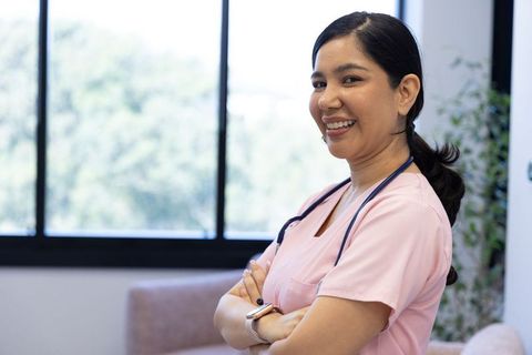Confident nurse smiling with arms crossed wearing pink scrubs and stethoscope in clinic