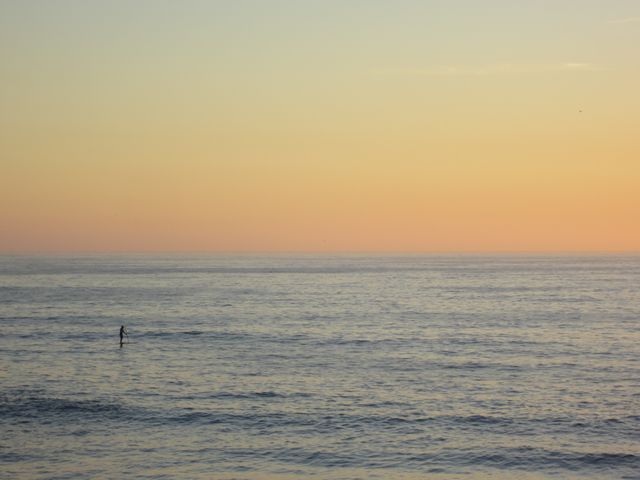This image depicts a person stand-up paddleboarding on the open water during a serene sunset. The sky transitions from shades of soft yellow to orange and pink, casting a peaceful glow over the calm ocean. Ideal for use in travel brochures, wellness blogs, or advertisements promoting outdoor activities and relaxation.