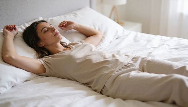 Woman relaxing on bed stretching arms in neutral loungewear morning calm minimalist bedroom