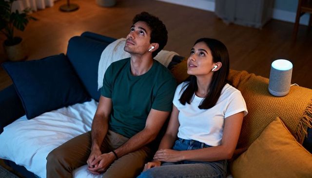 Young couple using smart speaker and wireless earbuds on cozy sofa at night