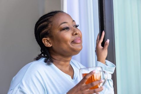 African american woman sipping tea at sliding glass door, morning calm in blue top