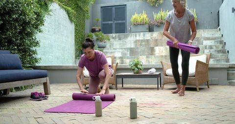 Mature women unrolling purple yoga mats on courtyard patio for outdoor wellness routine