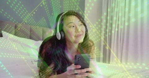 Relaxing asian woman listening on headphones and scrolling smartphone under holographic overlay