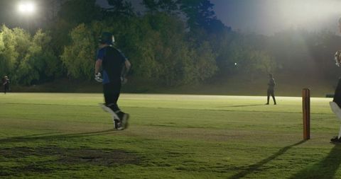 Teen cricket players sprinting between wickets under bright floodlights at night