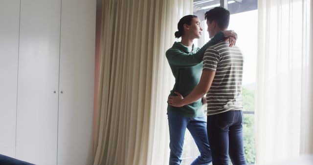 This close-up captures a young couple in a warm embrace, standing by a large window in a sunlit room. Ideal for use in marketing materials promoting relationships, love, or family themes. Can also be used in lifestyle blogs, articles about intimacy, or advertisements for home furnishings or real estate.