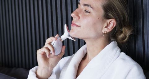 Woman using jawline sculpting facial device while relaxing in white robe
