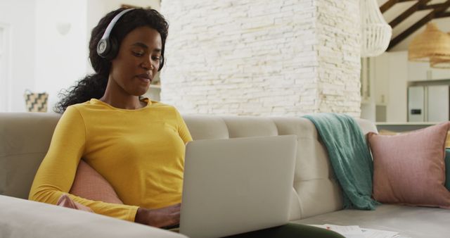 Image of happy african american woman on sofa with headphones using laptop. leisure and relax, spending free time with technology at home.