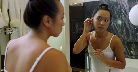 Asian Woman Reflecting in Mirror, Adjusting Earring in Bathroom