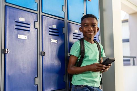 African american schoolboy leaning on blue lockers holding phone with backpack smiling