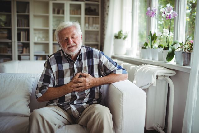 Senior man sitting on a white sofa, holding his chest in pain. The living room is well-lit with natural light coming through the windows. There are plants on the windowsill and bookshelves in the background. This image can be used in articles or advertisements related to health issues, elderly care, medical emergencies, and heart conditions.