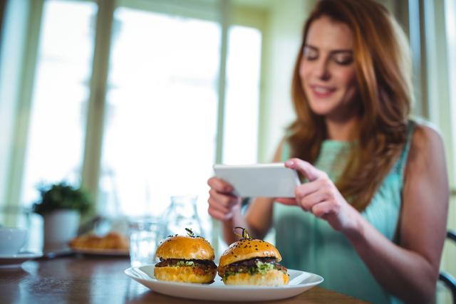Smiling woman clicking photo of burger from mobile phone in cafÃ©