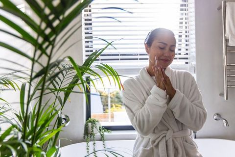 Asian woman applying skincare in sunlit bathroom robe beside freestanding tub and plants