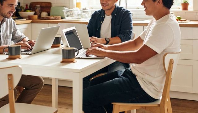Three Men Collaborating Remotely at Sunlit Kitchen Table with Laptops and Ceramic Mugs