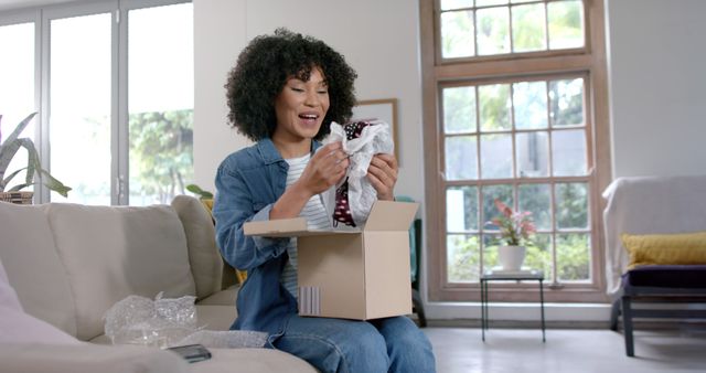 A woman with curly hair is joyfully unboxing a package while sitting on a couch in a well-lit living room. Ideal for illustrating concepts related to online shopping, home delivery, surprise gifts, and consumer satisfaction. Can be used in advertisements, blog posts, social media campaigns, and articles highlighting e-commerce, lifestyle, and seasonal promotions.