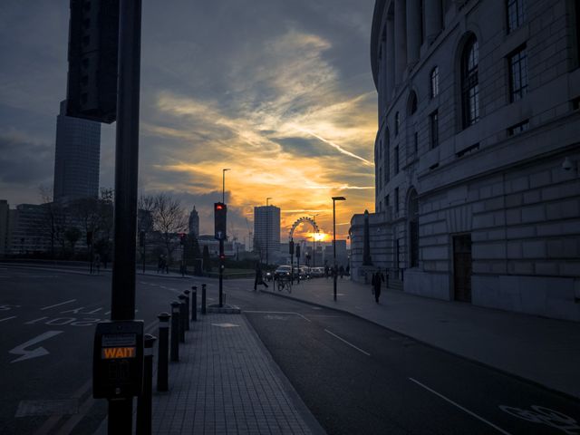This image captures a dramatic sunset over a city street with a Ferris wheel visible in the background. The architecture of the buildings, the streetlights, cars, and pedestrians create a typical urban atmosphere. Perfect for use in travel and tourism promotions, city lifestyle blogs, and urban architecture design projects.