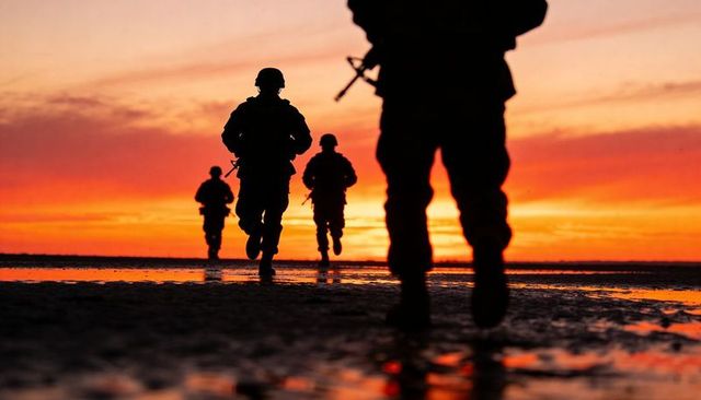 Running silhouetted soldiers on wet beach at fiery sunset with rifles and tactical gear
