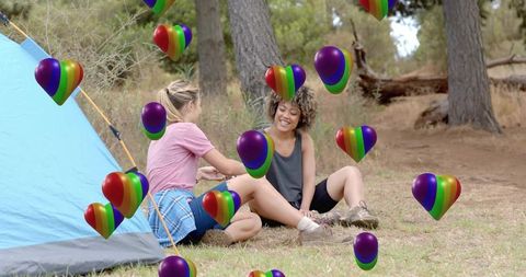 Friends camping laughing and chatting by tent with rainbow heart balloons in forest