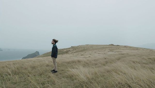 Windswept man standing on coastal headland facing misty ocean, contemplative seascape