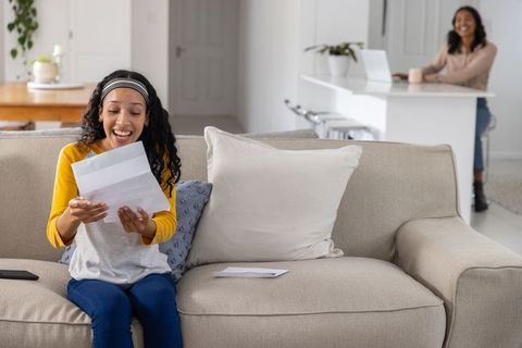 Teen daughter reading joyful acceptance letter while mother watching and smiling from kitchen
