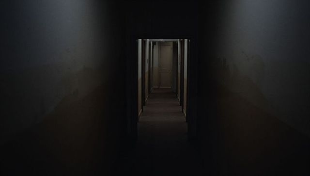 Deserted motel corridor leading toward closed beige door, dim moody worn interior