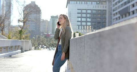 Woman leaning on bridge railing talking on smartphone holding coffee in sunlit city