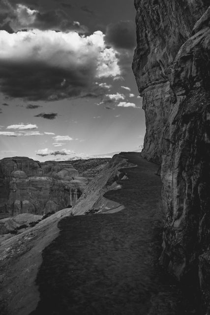 This dramatic black and white image showcases a desert trail winding between towering rock formations under a cloudy sky. Featuring strong contrasts and textured surfaces, it captures the rugged beauty of a natural landscape. Ideal for use in nature-inspired decor, travel articles, and outdoor adventure blogs.