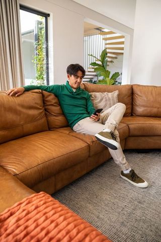 Young asian man relaxing on leather sofa using smartphone in modern open-plan living room