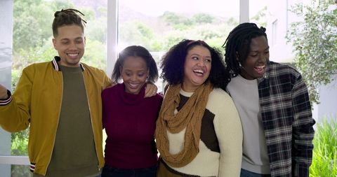 Diverse friends laughing and linking arms by floor-to-ceiling window with greenery