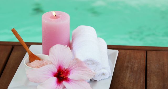 Perfect for illustrating spa and wellness concepts, this serene visual combines elements like a pink candle, rolled white towels, a fragrant hibiscus flower, and a bowl of salt on a wooden table by the calming poolside, ideal for promotional materials, spa websites, and relaxation-themed advertisements.