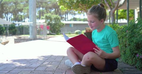 Young girl reading red notebook while sitting on brick courtyard near school playground