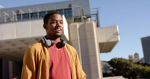 Standing young African American man wearing headphones on sunny urban campus, modern architecture