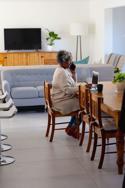 Vertical image of senior african american woman on phone, using laptop in dining room, copy space. Business, communication, working remotely, inclusivity and senior lifestyle concept.