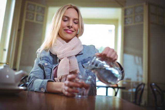 Woman sitting in a cafe, pouring water from a jug into a glass. She is smiling and wearing a scarf and denim jacket. Ideal for use in lifestyle blogs, hydration and wellness articles, cafe promotions, and casual dining advertisements.
