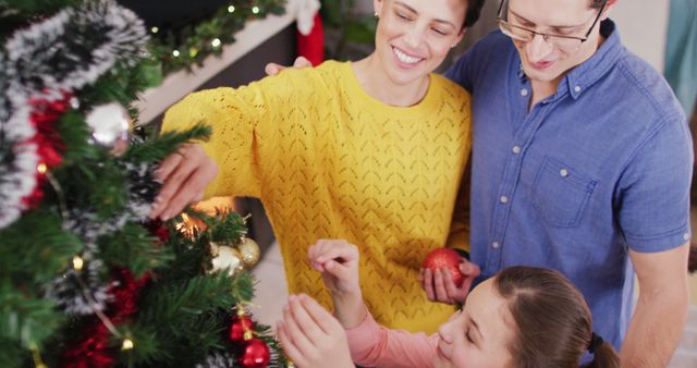 Family spending quality time decorating a Christmas tree together at home. Parents and child putting decorations on the tree, sharing smiles and warmth. Suitable for advertisements about family bonding during holiday season, festive promotions, and Christmas-themed content.