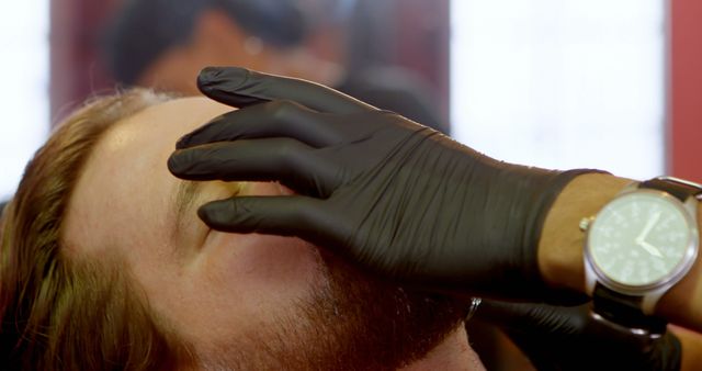 Close-up view of a professional barber grooming a male client's eyebrows using black gloves. The client has a beard and appears to be relaxed during the process. The barber is wearing a wristwatch that is visible near the client's face. This image is ideal for advertisements and content related to men's grooming, barbershops, personal care, and professional grooming services.