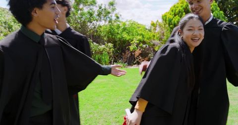 Diverse students celebrating graduation on green lawn wearing black academic gowns