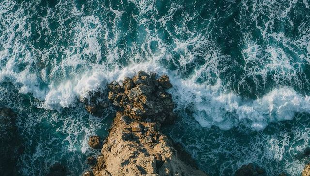 Aerial teal ocean waves crashing on jagged rocky promontory with swirling white foam