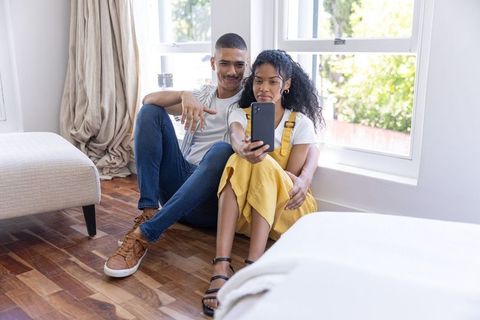 Sunlit african american couple sitting on bedroom floor taking selfie, casual home moment