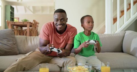 African American father and son playing video games on couch with snacks and orange juice
