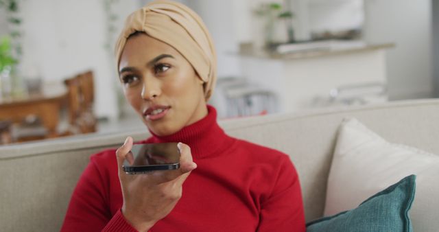 Image of happy biracial woman in hijab sitting on sofa and having call. Lifestyle, spending free time at home with technology concept.