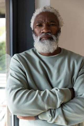 African american senior man standing by window with arms crossed wearing sage sweatshirt