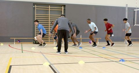 Diverse male athletes performing lateral agility drills with coach in school gymnasium