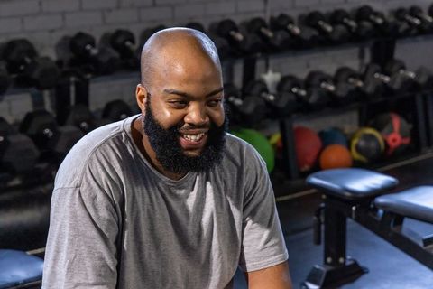 Smiling african american man resting in gym among dumbbells, medicine balls and benches