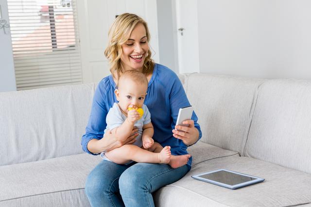 Mother sitting on couch, holding baby boy and using mobile phone, likely for communication or leisure. Baby chewing on toy, both appearing happy and engaged. Suitable for concepts of modern parenting, technology in family life, staying connected, and daily routines at home.