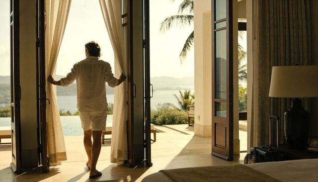 Man standing at villa doors holding curtains gazing at ocean and infinity pool