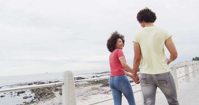 This image depicts a couple holding hands and walking by the beach, with the ocean in the background and a cloudy sky overhead. They seem happy and relaxed, dressed in casual clothing. This photo could be used for advertisements related to travel, romance, lifestyle blogs, or articles focused on relationships and leisure activities.
