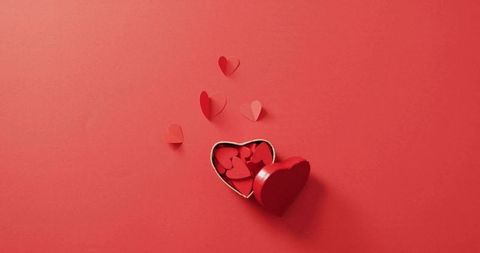 Red heart-shaped gift box spilling paper hearts on monochrome red backdrop for romance