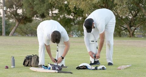 Cricket players fastening batting pads on grass field before match, teamwork preparation