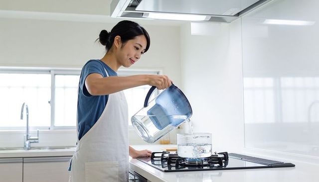 Woman pouring water from pitcher into glass pot on gas stove in minimalist modern kitchen